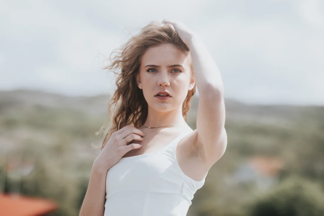 Woman in a white tank top outdoors, one hand in her hair, looking into the camera